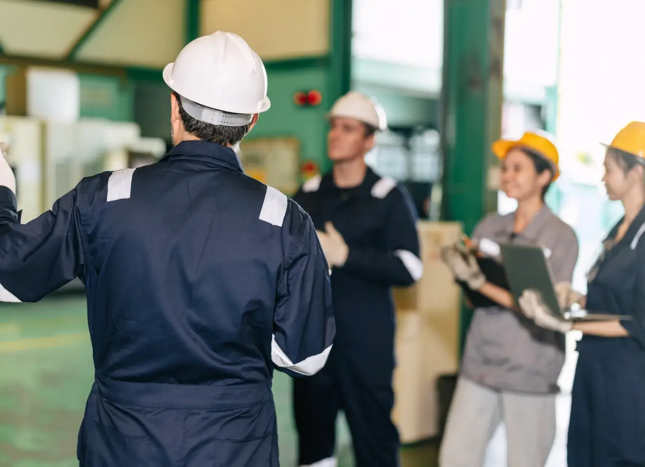 Supervisor in a white hard hat addressing a group of factory workers wearing hard hats and holding clipboards on an industrial floor.