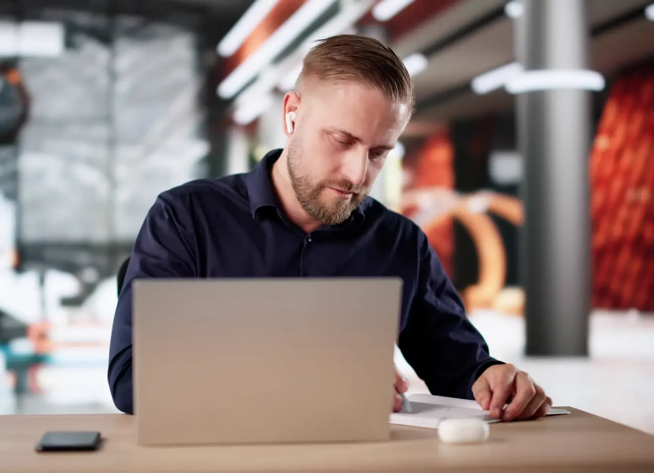 Man in a dark shirt reviewing documents at a desk with a laptop and smartphone in a modern office setting.