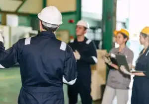 Supervisor in a white hard hat addressing a group of factory workers wearing hard hats and holding clipboards on an industrial floor.